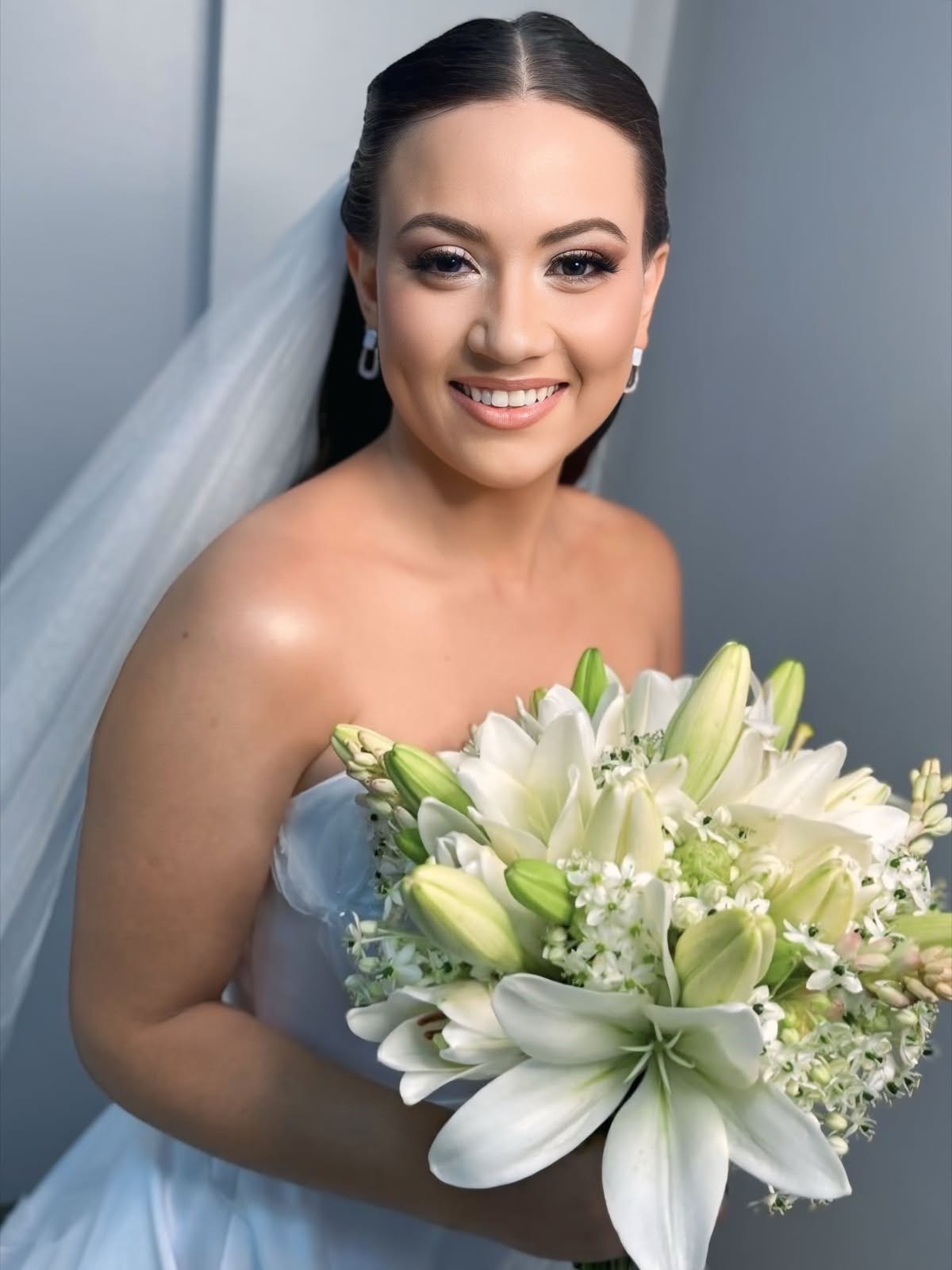 Noiva sorrindo com véu e buquê de lírios brancos, maquiagem e penteado impecáveis