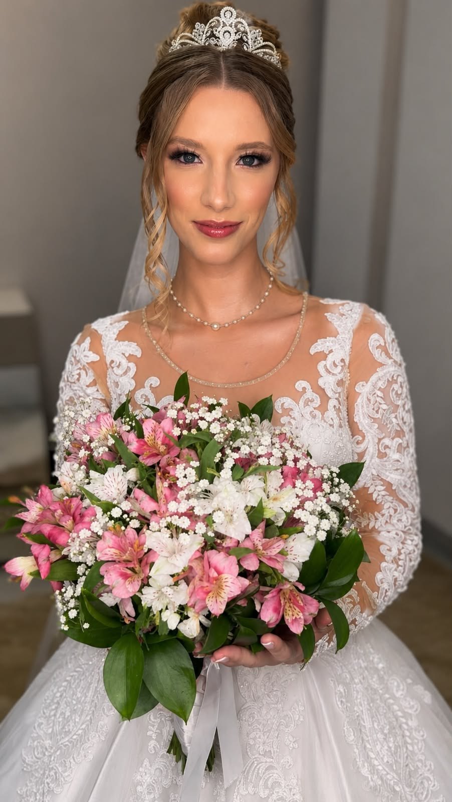 Noiva com tiara, véu e vestido de renda, segurando buquê de astromélias rosas e brancas, maquiagem e penteado delicados