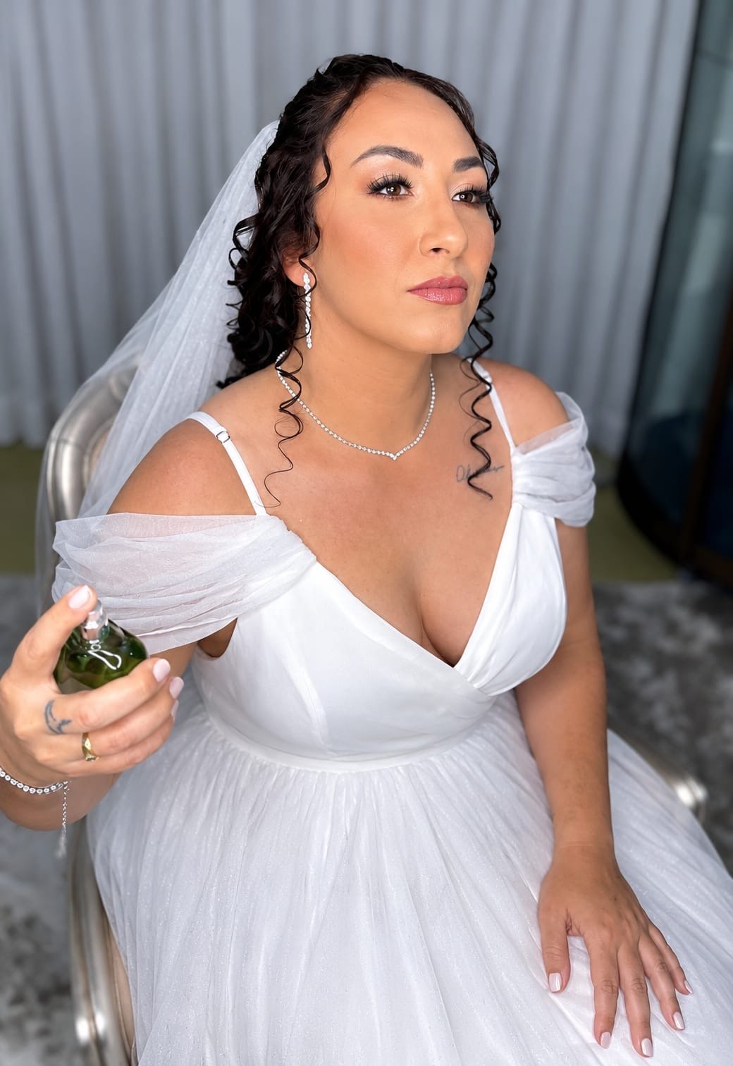Noiva com véu e vestido de tule sentada, segurando um perfume, maquiagem suave e cachos soltos