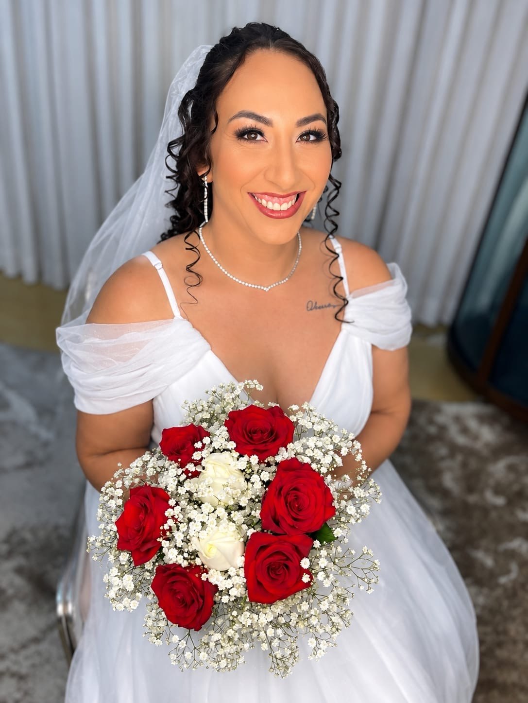 Noiva sorrindo com buquê de rosas vermelhas e brancas, véu e colar de brilhantes